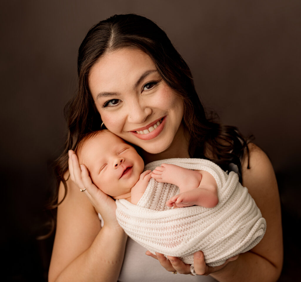 new mother in white tank top holds newborn baby boy wrapped in white blanket 