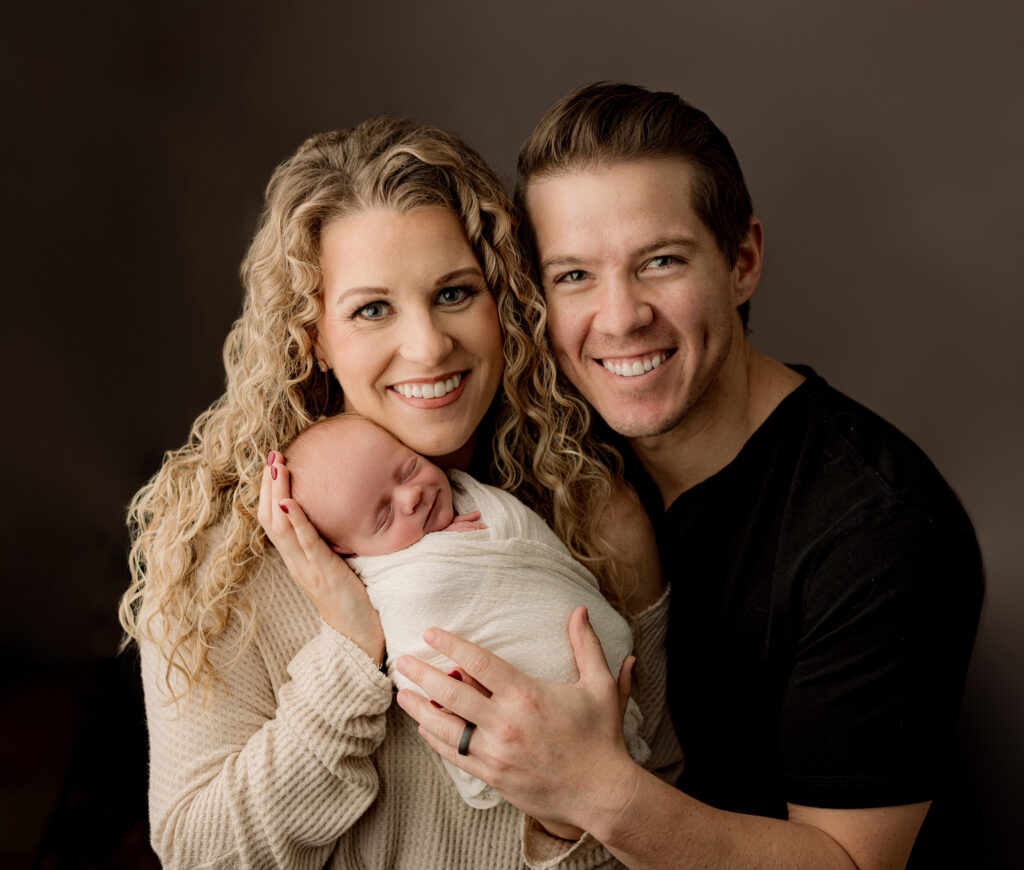 new mother with blonde curly hair holds newborn son wrapped in white blanket with dad hugging them 