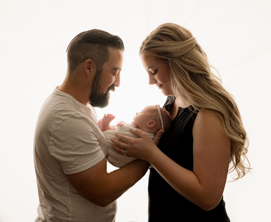 mom and dad look down at baby girl wrapped in white blanket at Austin newborn photography studio