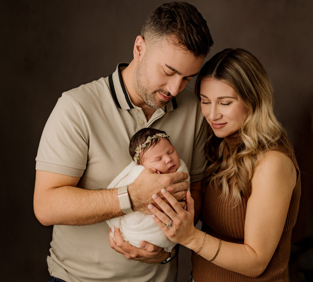 dad and mom hold newborn daughter wrapped in white blanket while looking down at her 