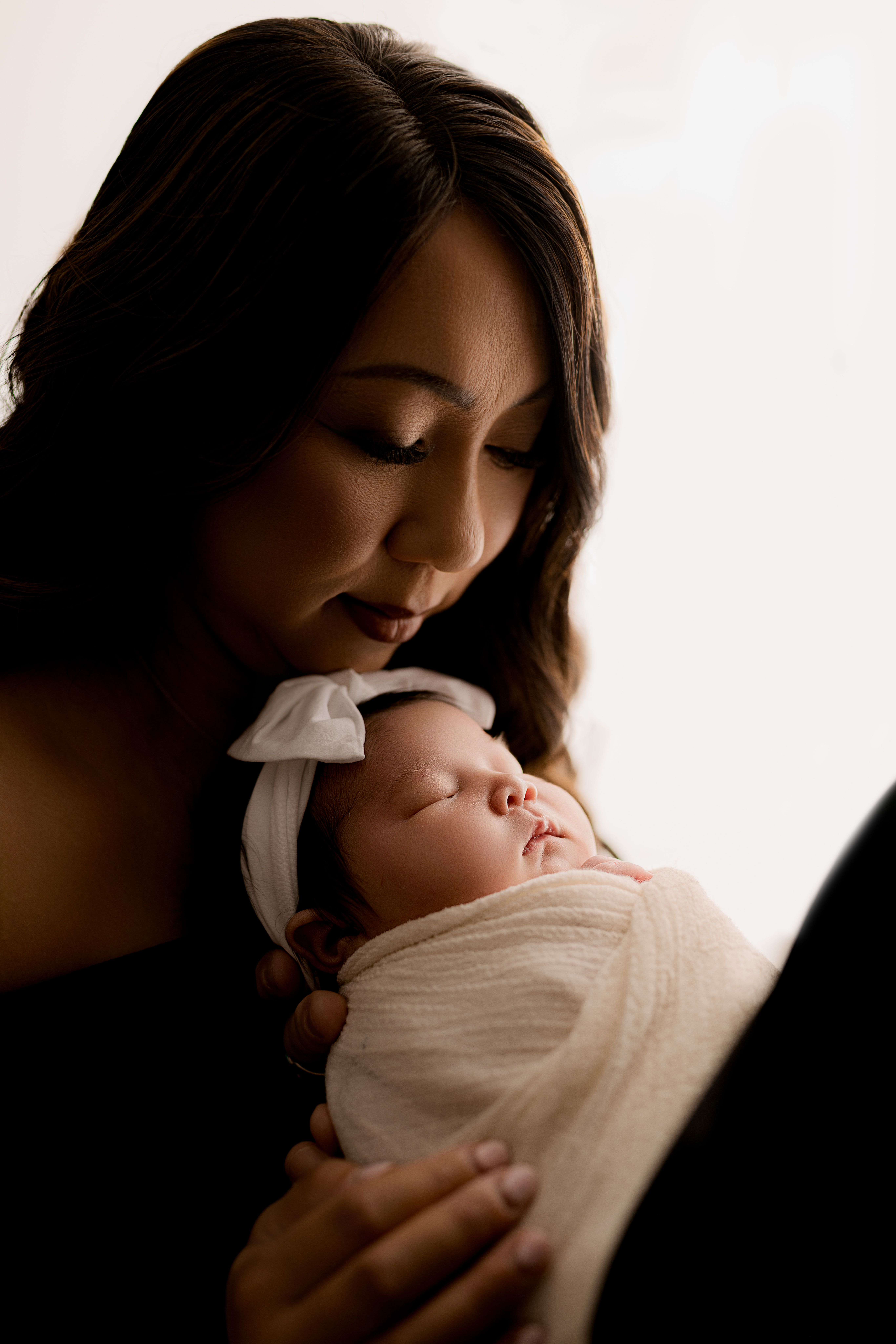 Mom in black dress holds and looks down at baby in white blanket with white bow on her head