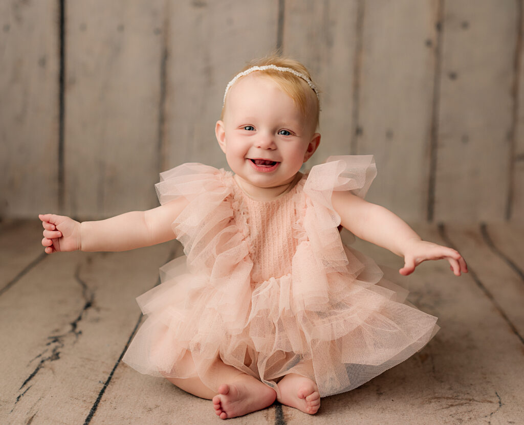 6 month old in pink dress sits with hands out and smiles at camera