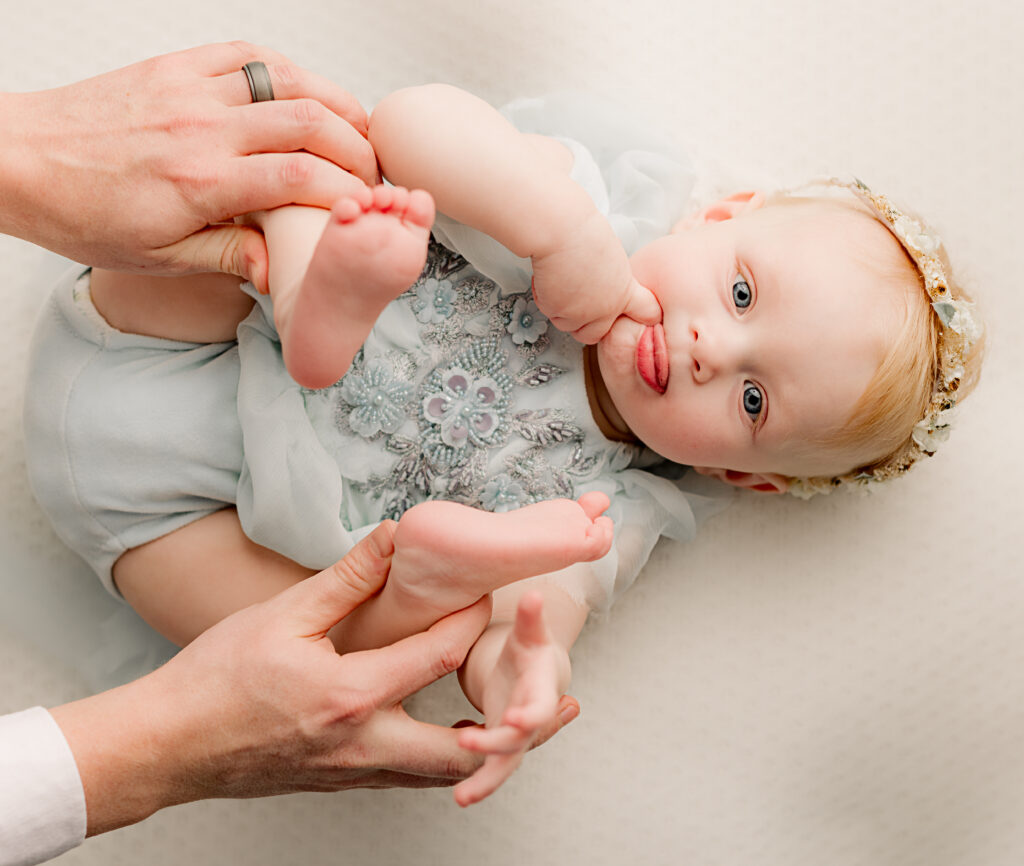 6 month old baby girl look at camera with finger in her month and tongue out while her dad holds her feet 