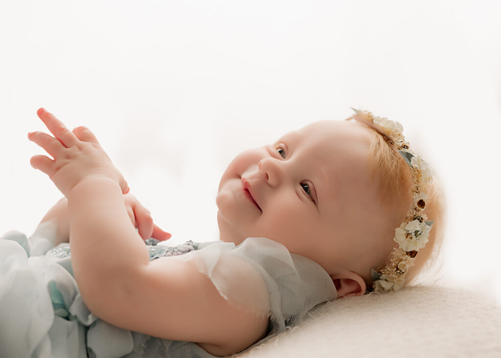 Baby in blue dress llooks up and smiles on white backlit backdrop 