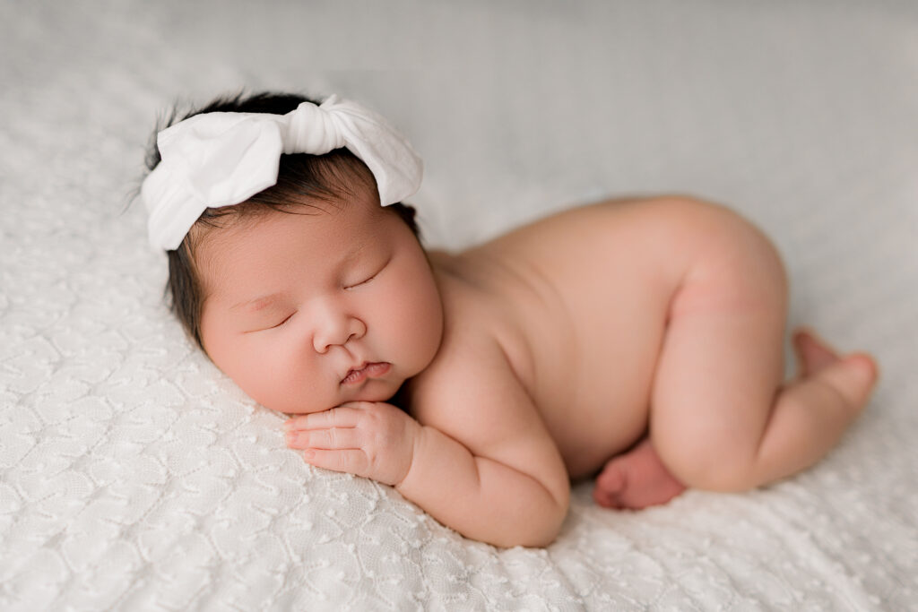 Baby laying on white backdrop with white bow in her long black hair 
