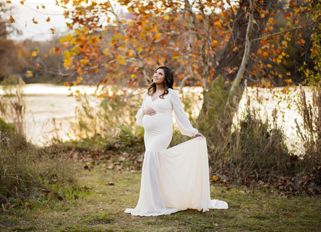 pregnant mother stands in a white dress holding one hand on her belly and the other holding the dress out while she looks up at the beautiful fall sky in feild with river and fall colored leaves behind her 