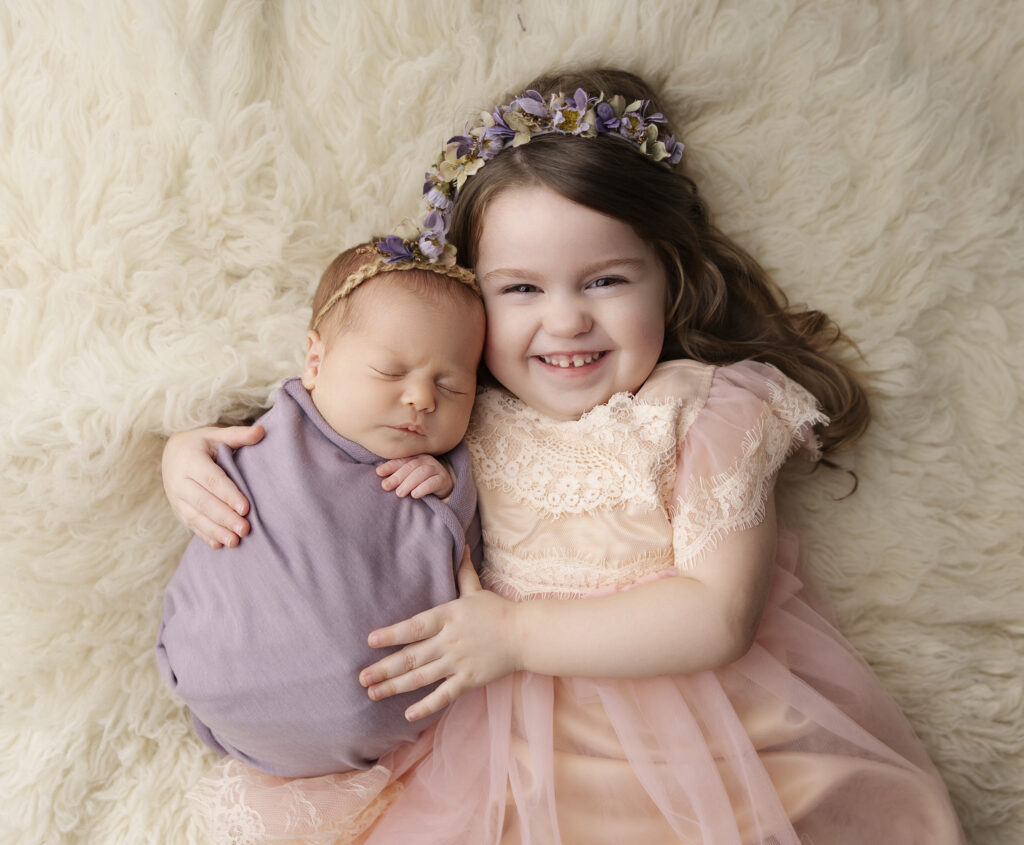 Baby wrapped in purple wrap held by smiling big sister in pink lace dress with matching floral purple headbands on in Austin Texas newborn phtography studio