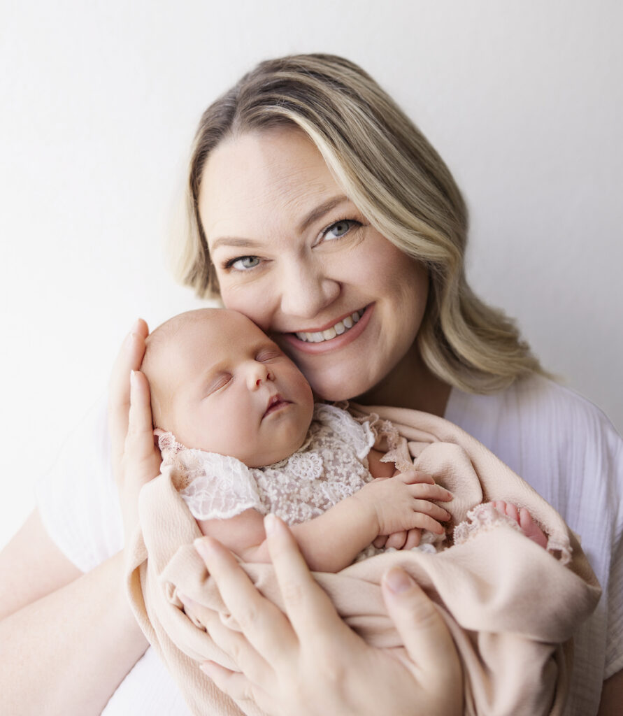 Mother smiles while holding her newborn baby girl wrapped in pink wrap up to her cheek on white backdrop 