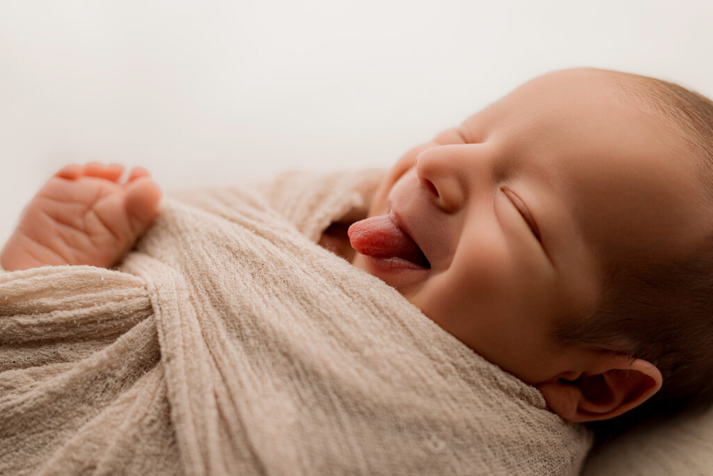 Baby boy sticks out tongue wrapped in tan wrap with foot sticking out 