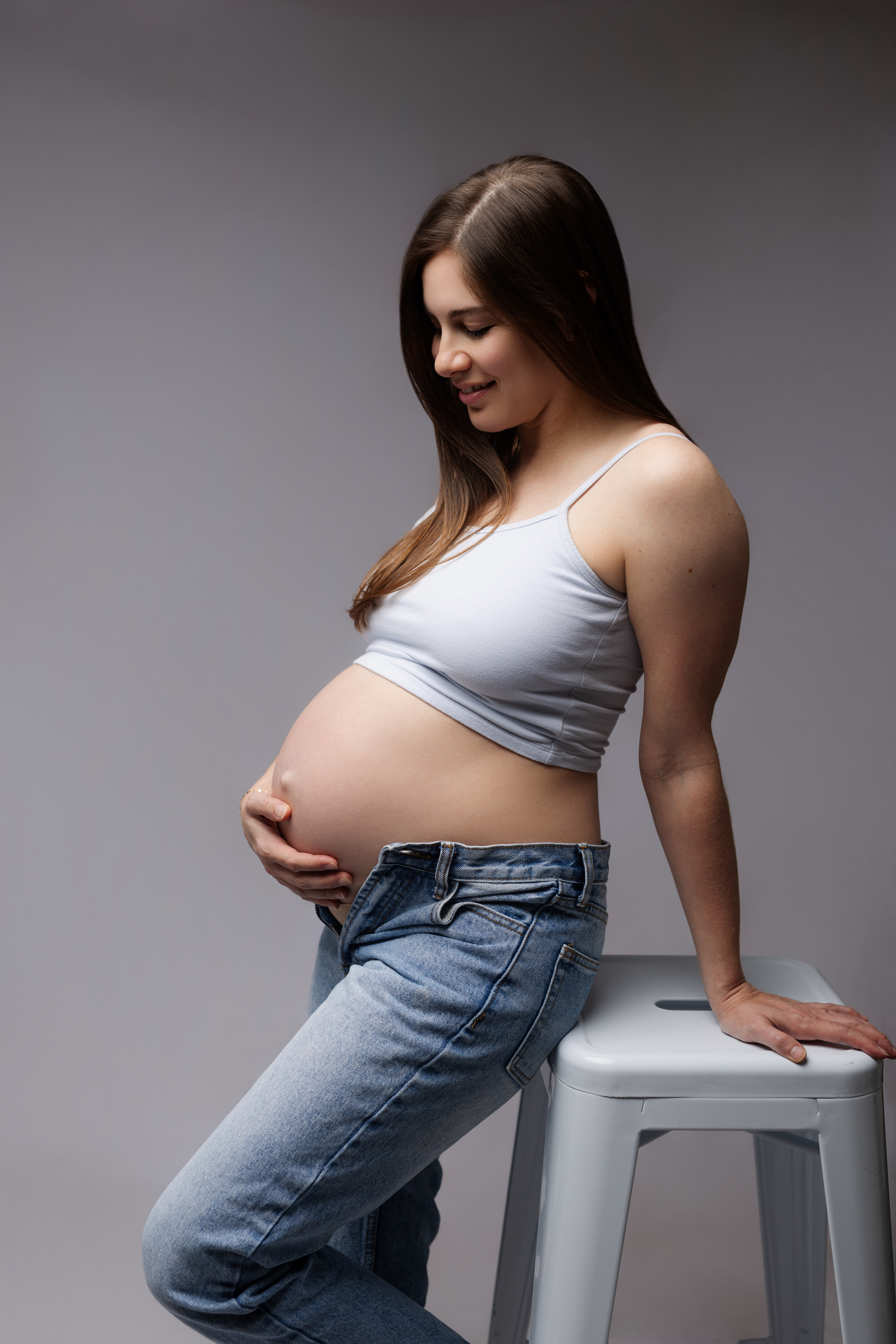Pregnant mother in white tank top and jeans poses. by leaning on white stool while looking down at her baby bump at a maternity photography studio in Austin